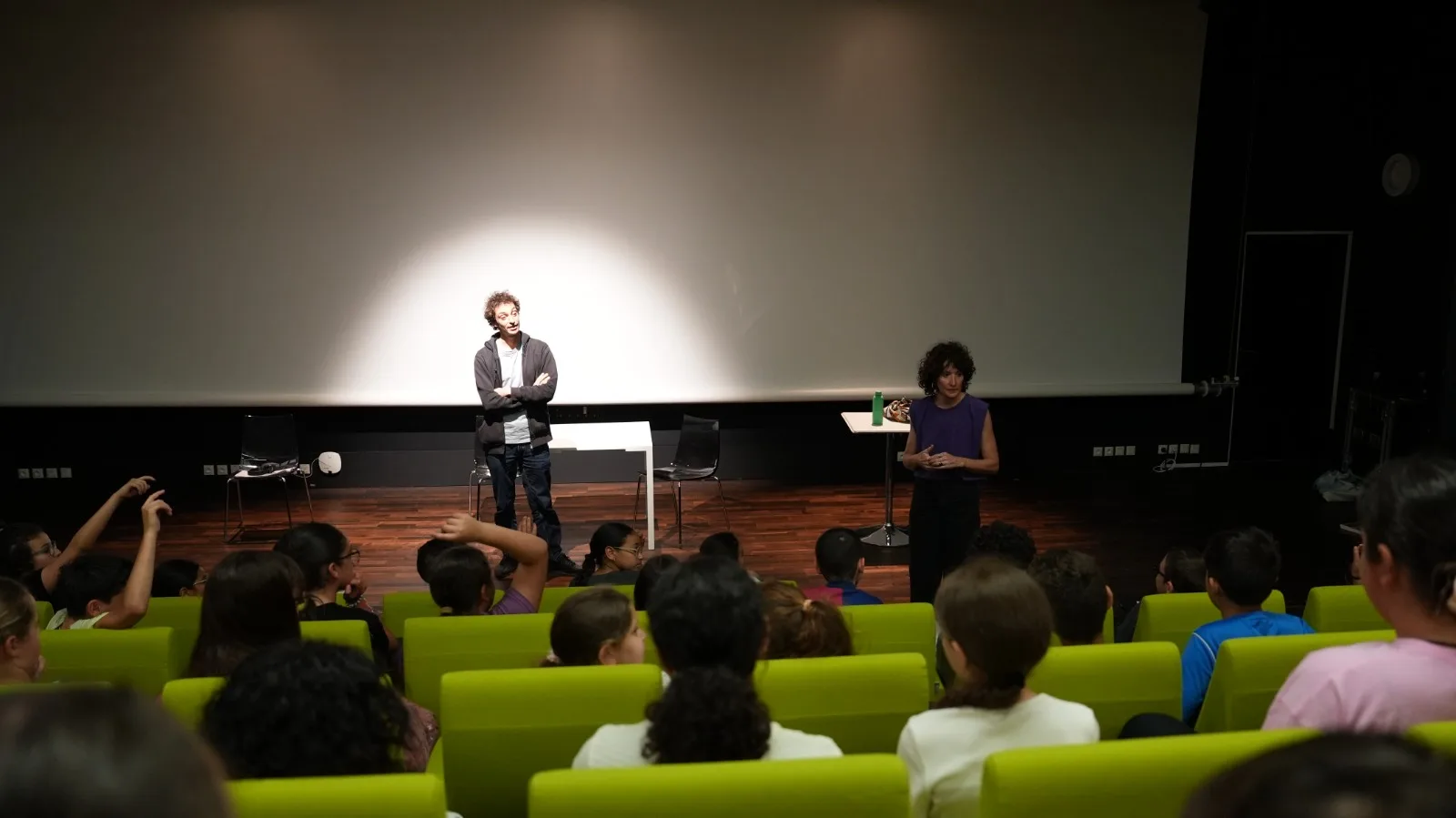 Samy et Béatrice sur scène devant une salle pleine d'enfants sur sièges verts