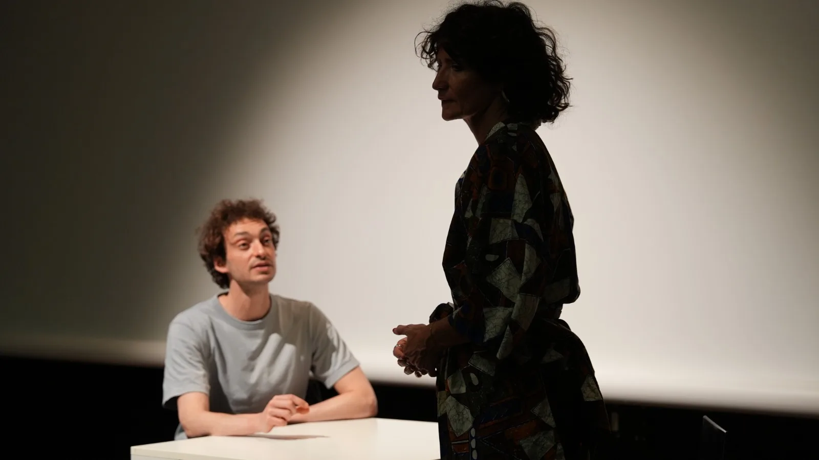 Samy assis à la table, Béatrice debout de dos, scène dramatique en contre-jour